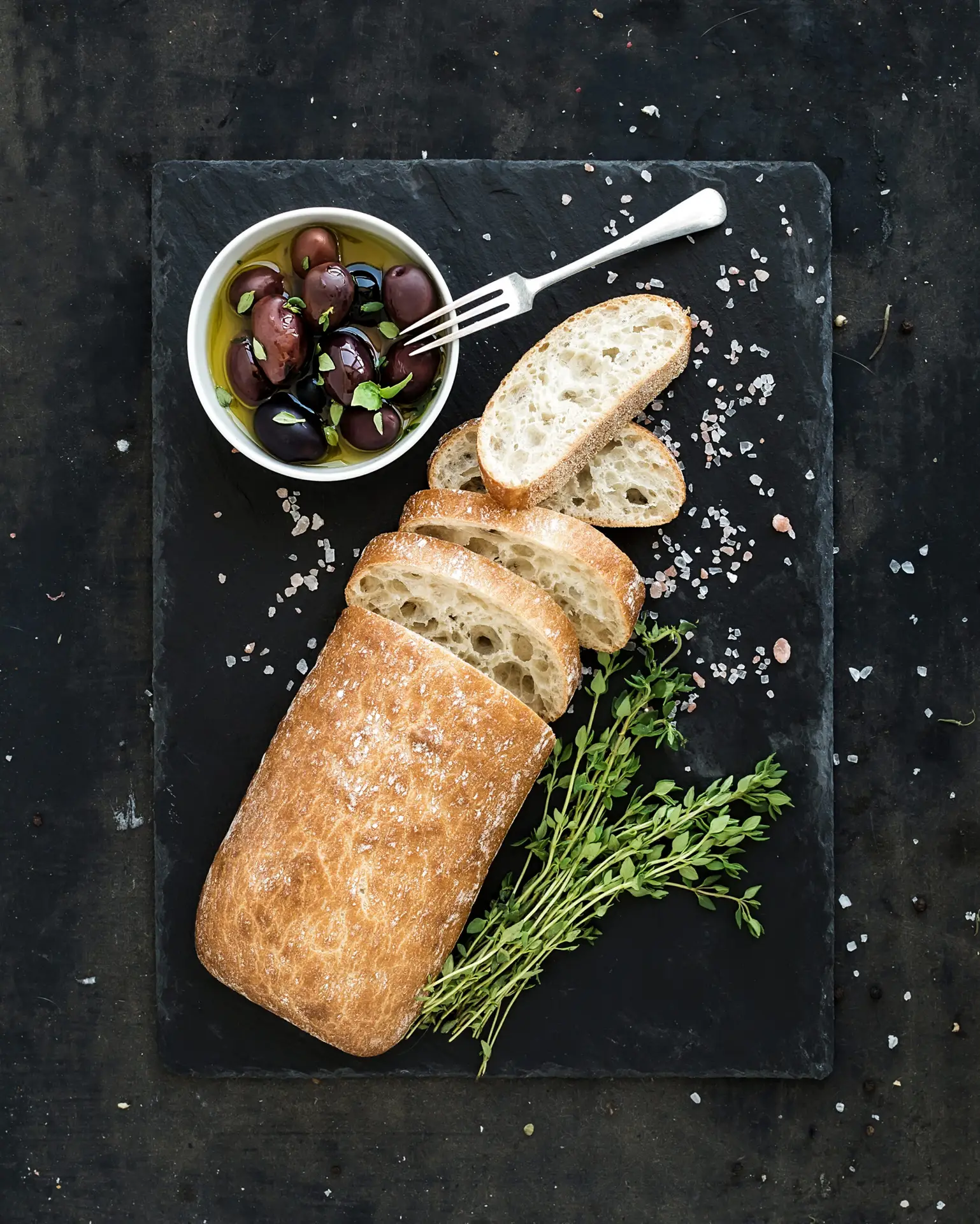 Italian ciabatta bread cut in slices on wooden chopping board with herbs, garlic and olives over dark grunge backdrop, top view